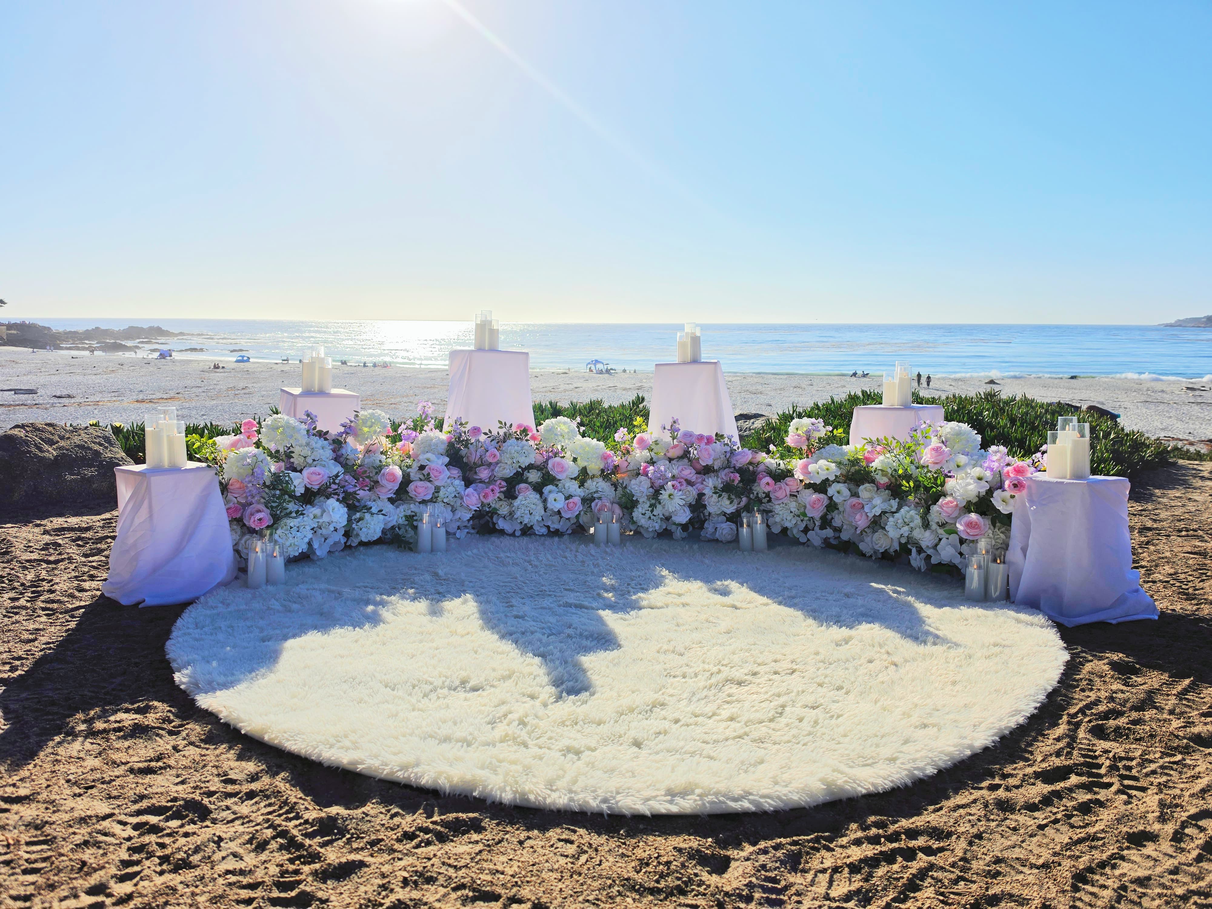 The Enchanted Romance Package on the sand at Carmel Beach, with a sweeping arc of blush pink, white, and lavender florals, candlelit white draped pedestals, and a fluffy white rug facing the Pacific