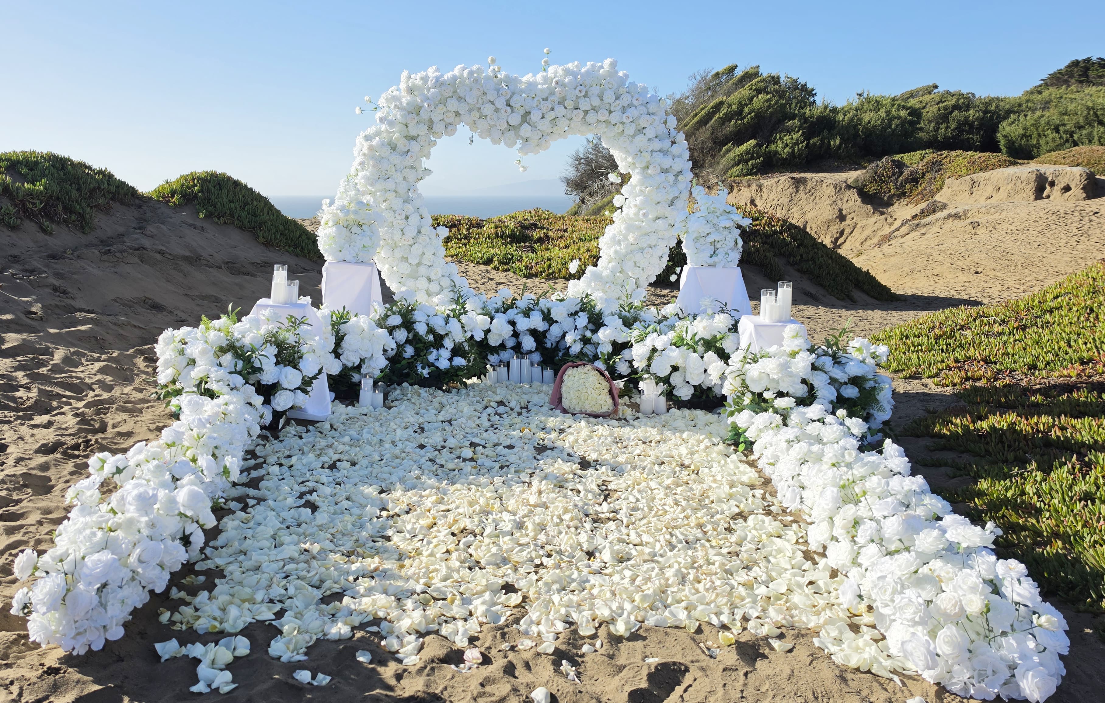 The Eternal Bloom – front view of the all-white floral heart arch above ten lush grounded floral arrangements, with a 20,000 rose petal walkway framed by four flower rows and a white rose bouquet at the center, at Fort Funston, San Francisco