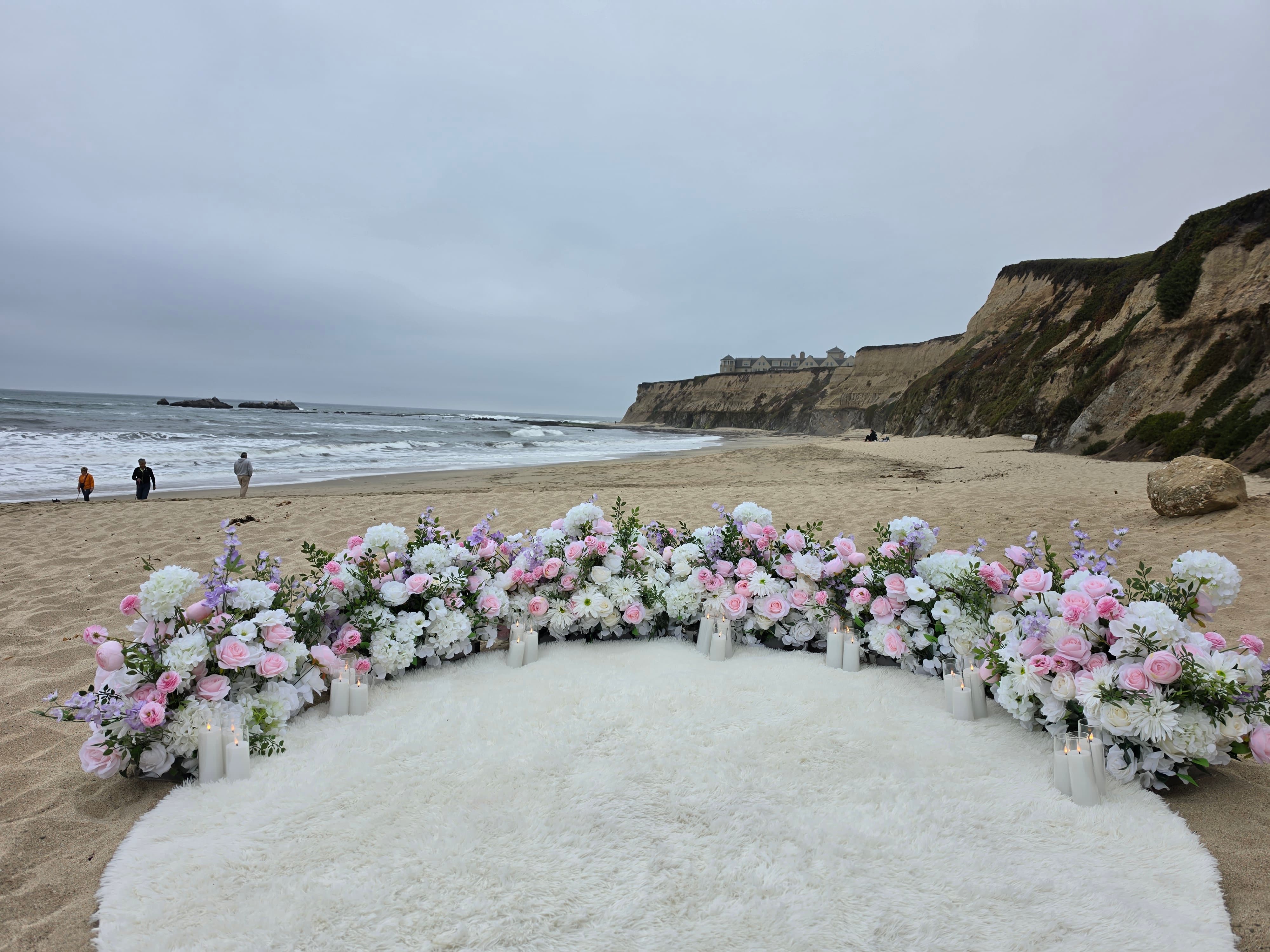 The Intimate Promise Package on Manhattan Beach, El Granada, with a crescent of blush pink, white, and lavender grounded florals, pillar candles, and a fluffy white rug on the sand above the Pacific