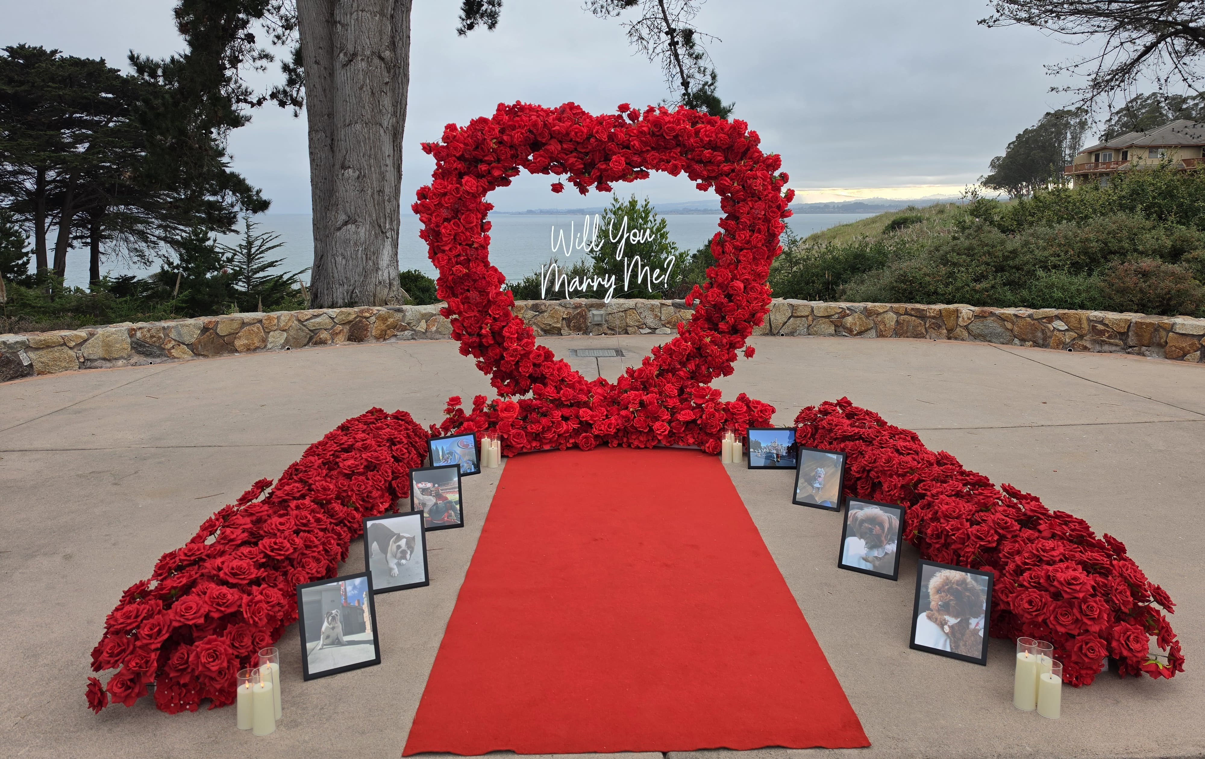 The Ever After Package on the Bluff terrace at Seascape Beach Resort, with a red rose heart arch and a glowing Will You Marry Me neon sign overlooking Monterey Bay, and a red aisle runner lined with red roses, framed photos, and pillar candles