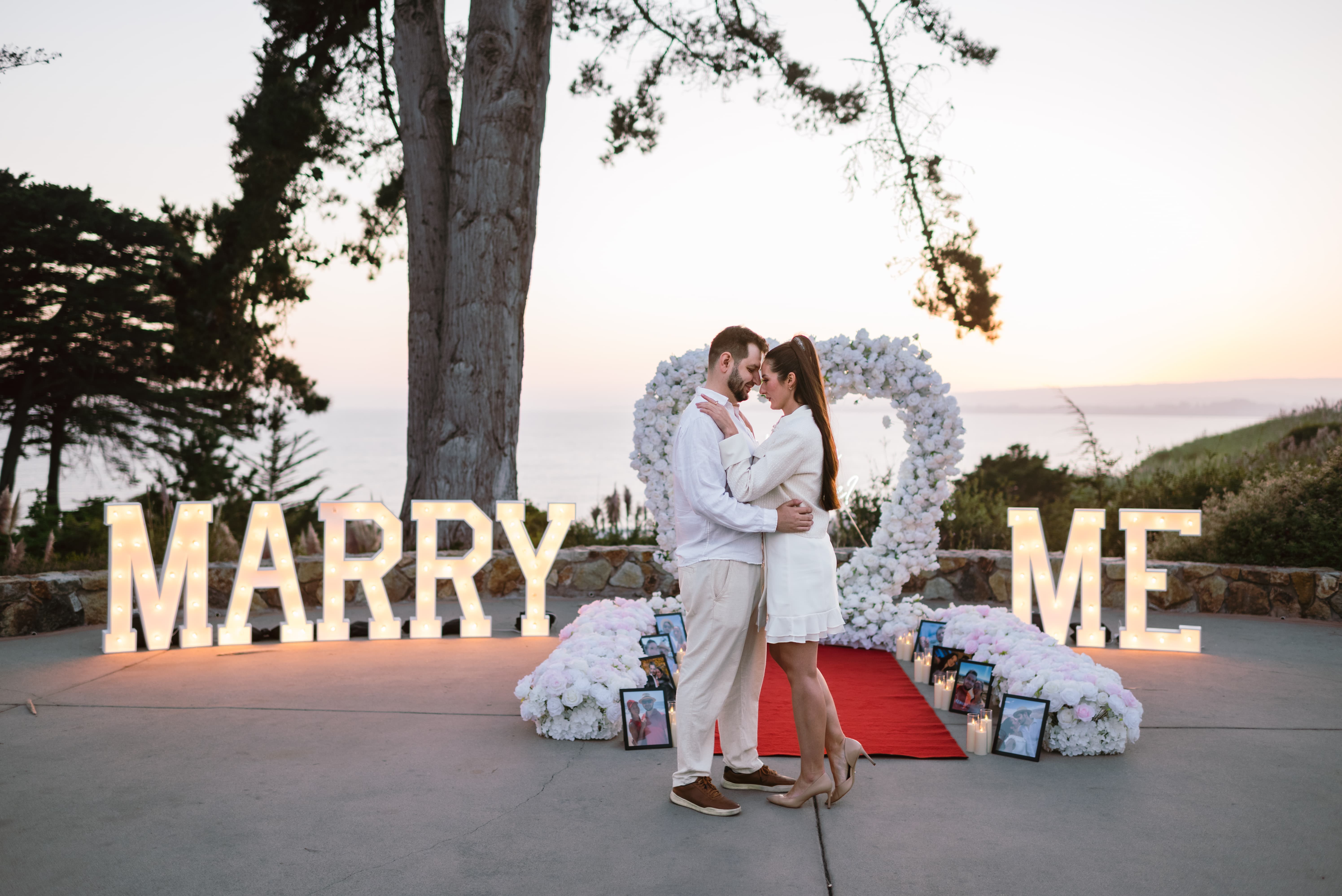 Couple embracing at sunset on the Bluff terrace at Seascape Beach Resort in front of a white floral heart arch, glowing MARRY ME marquee letters, and a white and blush floral aisleway above Monterey Bay
