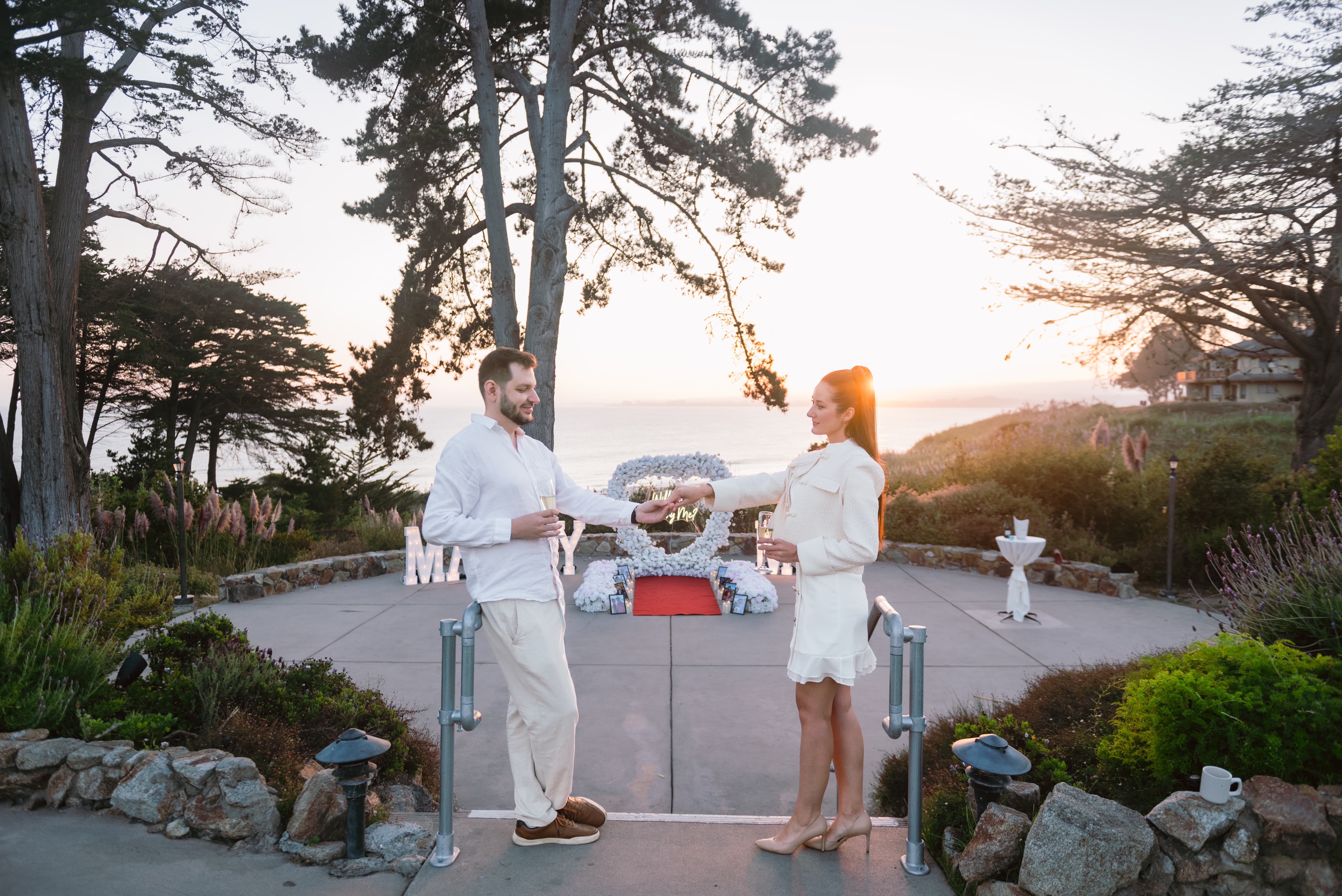 Newly engaged couple toasting with champagne on the Bluff terrace at Seascape Beach Resort, with the white floral heart arch, marquee letters, and the sun setting over Monterey Bay behind them