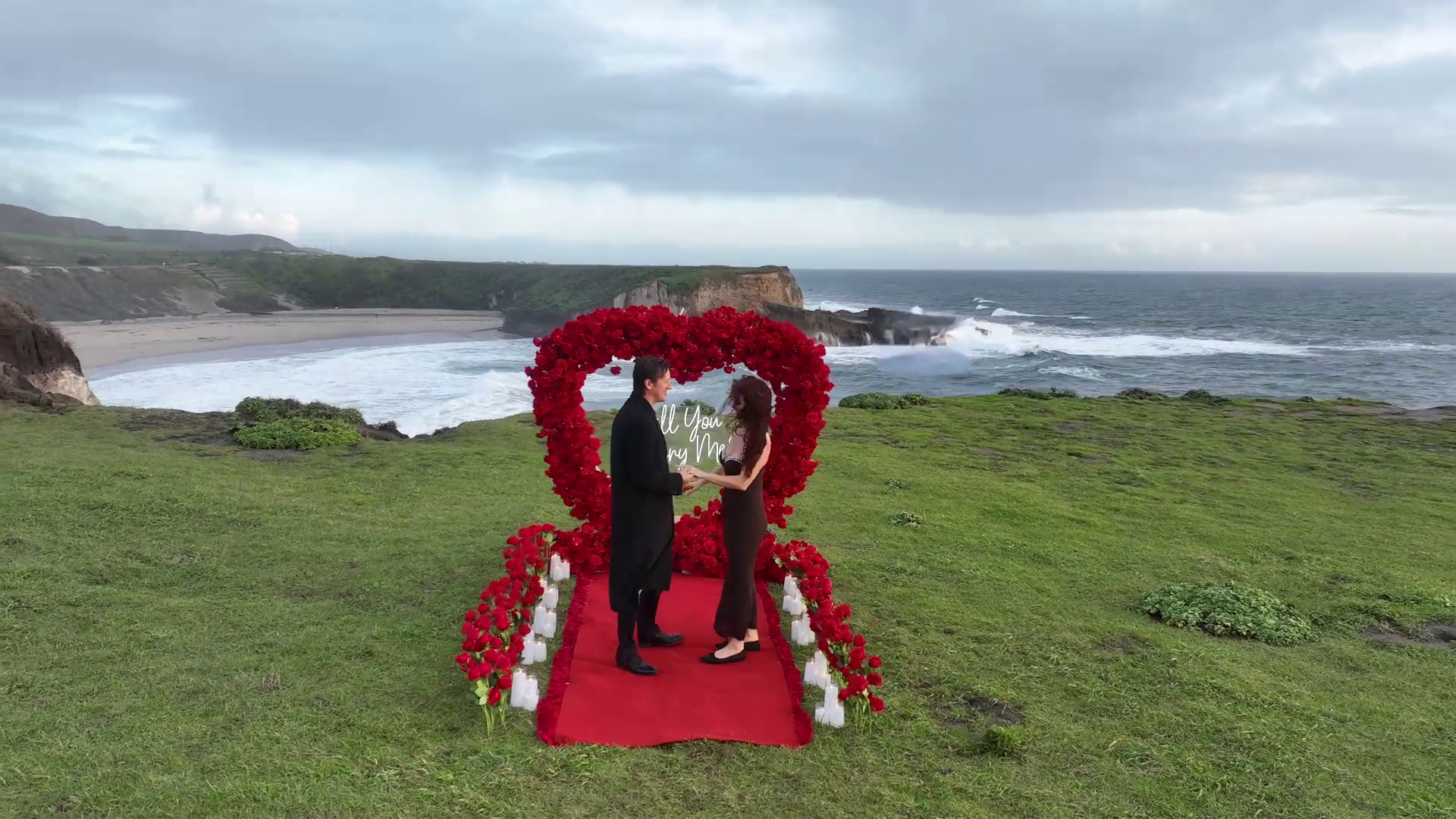 Elaborate floral heart arch proposal setup on a grassy California clifftop